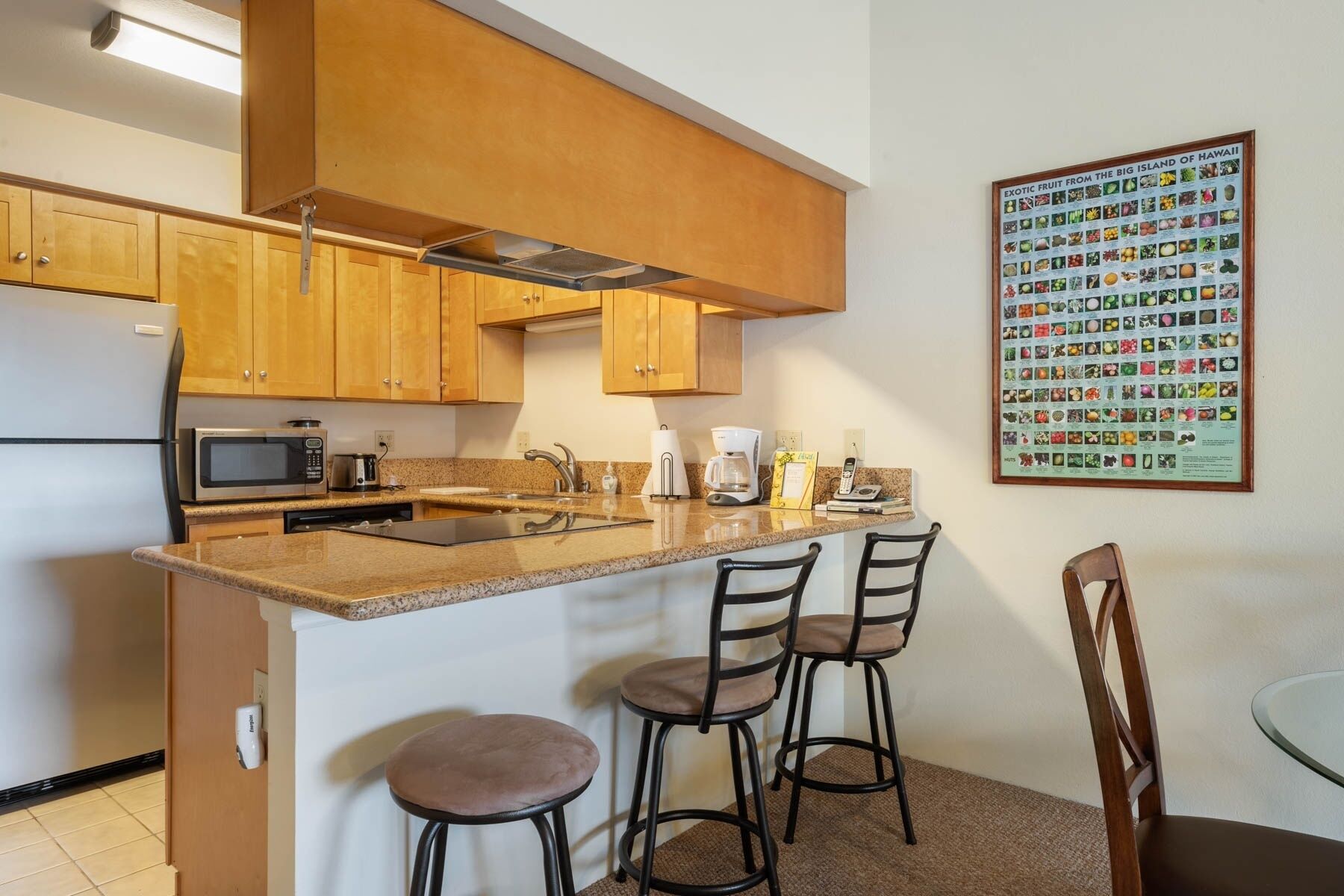 A modern kitchen with wooden cabinets, a granite countertop, a microwave, a coffee maker, and a framed poster of exotic fruits.