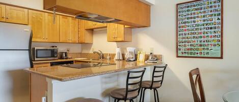 A modern kitchen with wooden cabinets, a granite countertop, a microwave, a coffee maker, and a framed poster of exotic fruits.