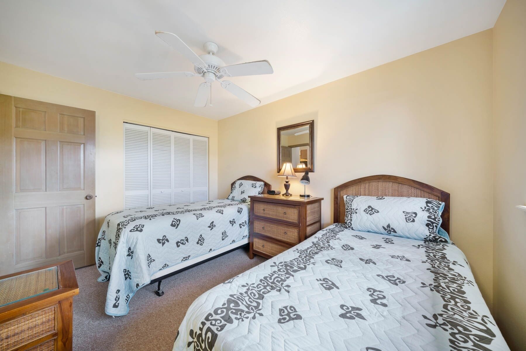 A bedroom with two beds, a ceiling fan, a bedside table with a lamp, and a mirror on the wall.