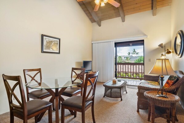 A dining area with a glass-top table and wooden chairs, a small round table with a wicker chair, a sofa with a lamp, and a view of the outdoors through a large window.