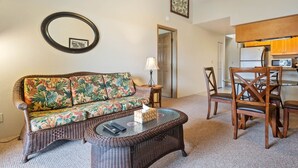 A living room with a sofa, a coffee table, a dining table with chairs, a mirror, and a kitchen area with appliances.