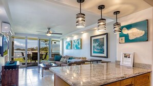A spacious kitchen with a granite countertop, wooden cabinets, and pendant lighting. There is a dining area with a table and chairs, and a large window offering a view of the outdoors. - Royal Mauian by Coldwell Banker Island Vacations (Kihei)