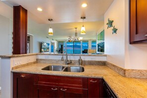 A kitchen with granite countertops, a double sink, and wooden cabinets. There are pendant lights above the sink and a large mirror on the wall. - Royal Mauian by Coldwell Banker Island Vacations (Kihei)