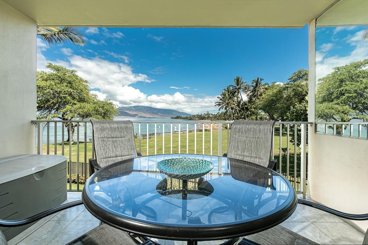 Un balcon doté d’une table et de chaises en verre, offrant une vue sur une zone verdoyante et un plan d’eau, avec des montagnes à l’horizon.