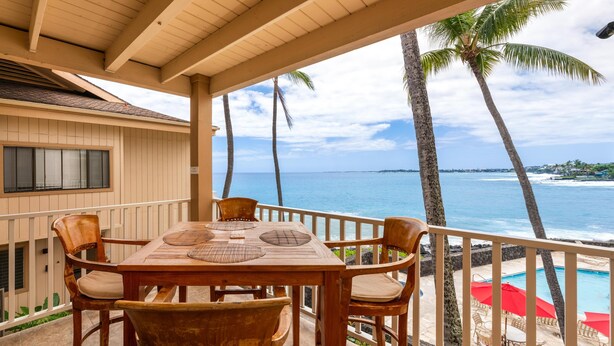 A wooden table with chairs on a balcony overlooking a pool and ocean. - Big Island Sea Village 3212 2 Bedroom Condo (Kailua-Kona)