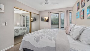 A bedroom with a bed, a ceiling fan, a TV, and a door leading to another room. - Big Island Sea Village 3212 2 Bedroom Condo (Kailua-Kona)