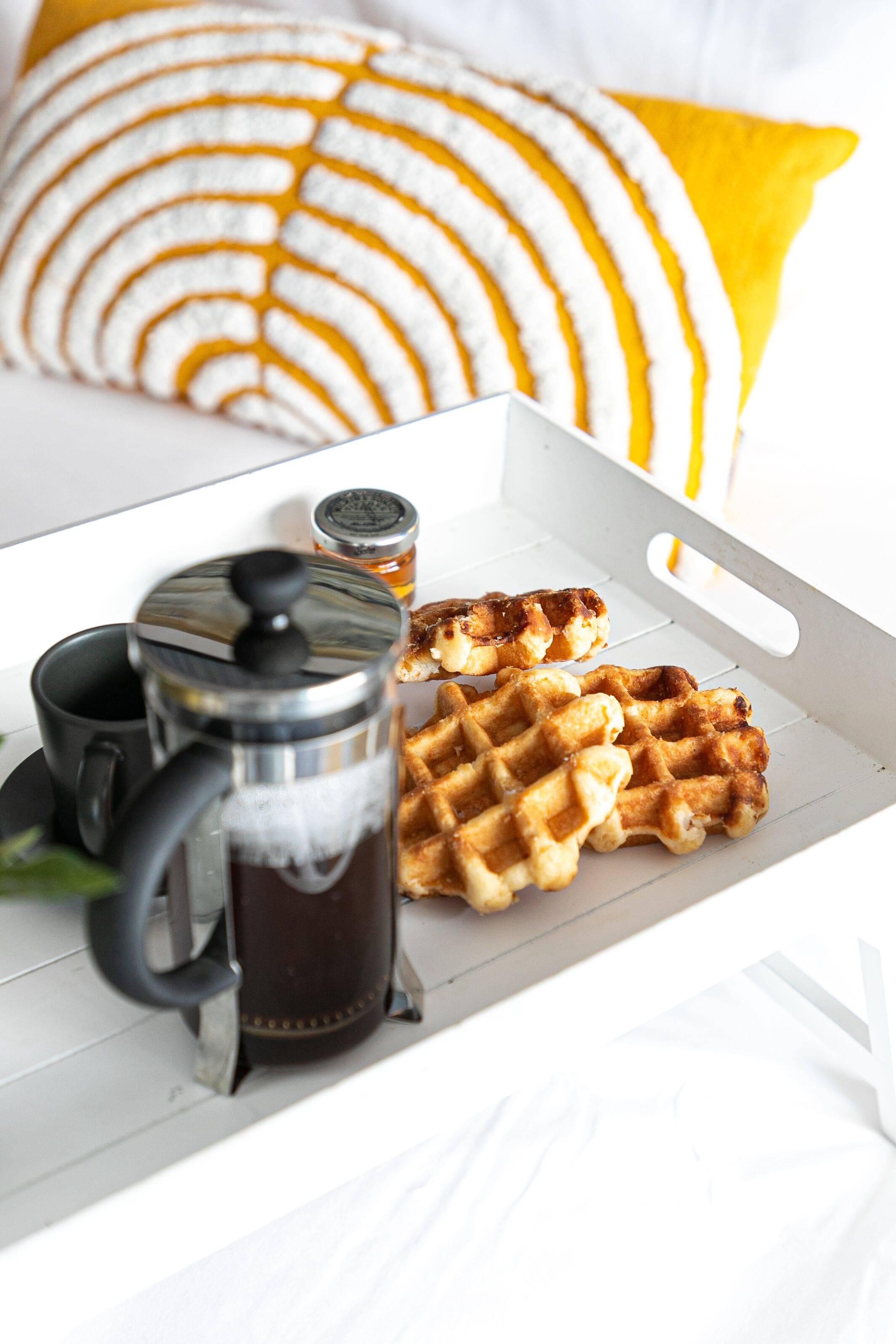 A tray with waffles, a coffee pot, and a cup on a white surface with a yellow pillow in the background.