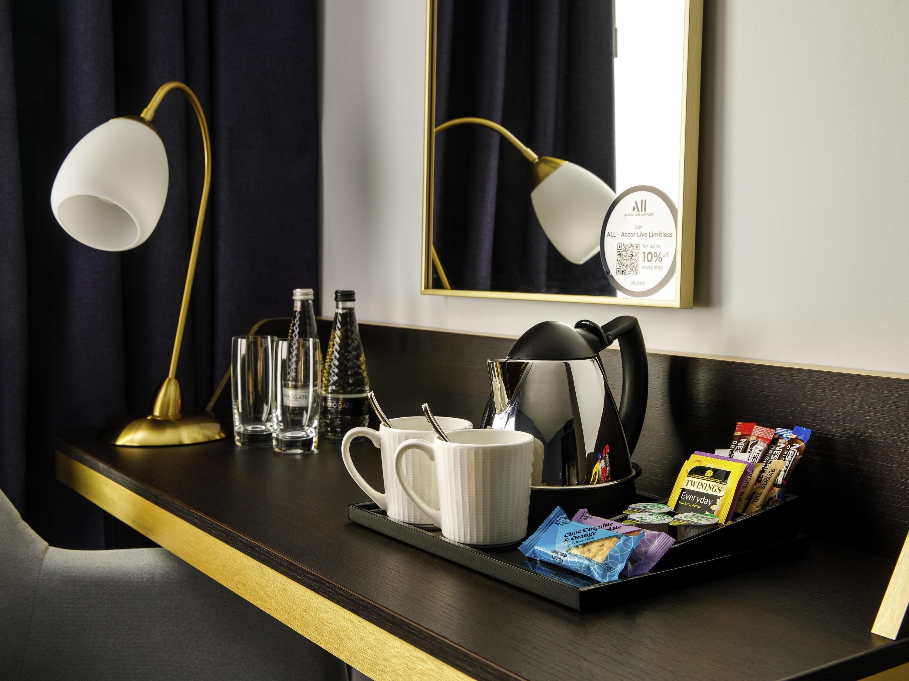 Hotellihuoneessa on tumma puinen työpöytä, valkoinen muki, tarjotinalus, jossa on Twinings-teepusseja, ja musta vedenkeitin.