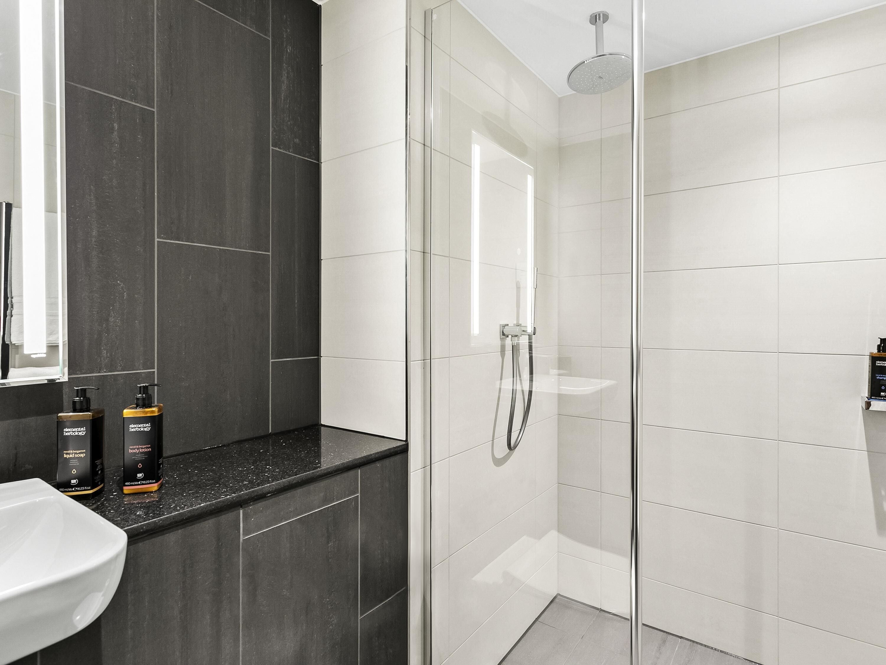 A modern bathroom with a glass-enclosed shower, a white sink, and dark tiles.