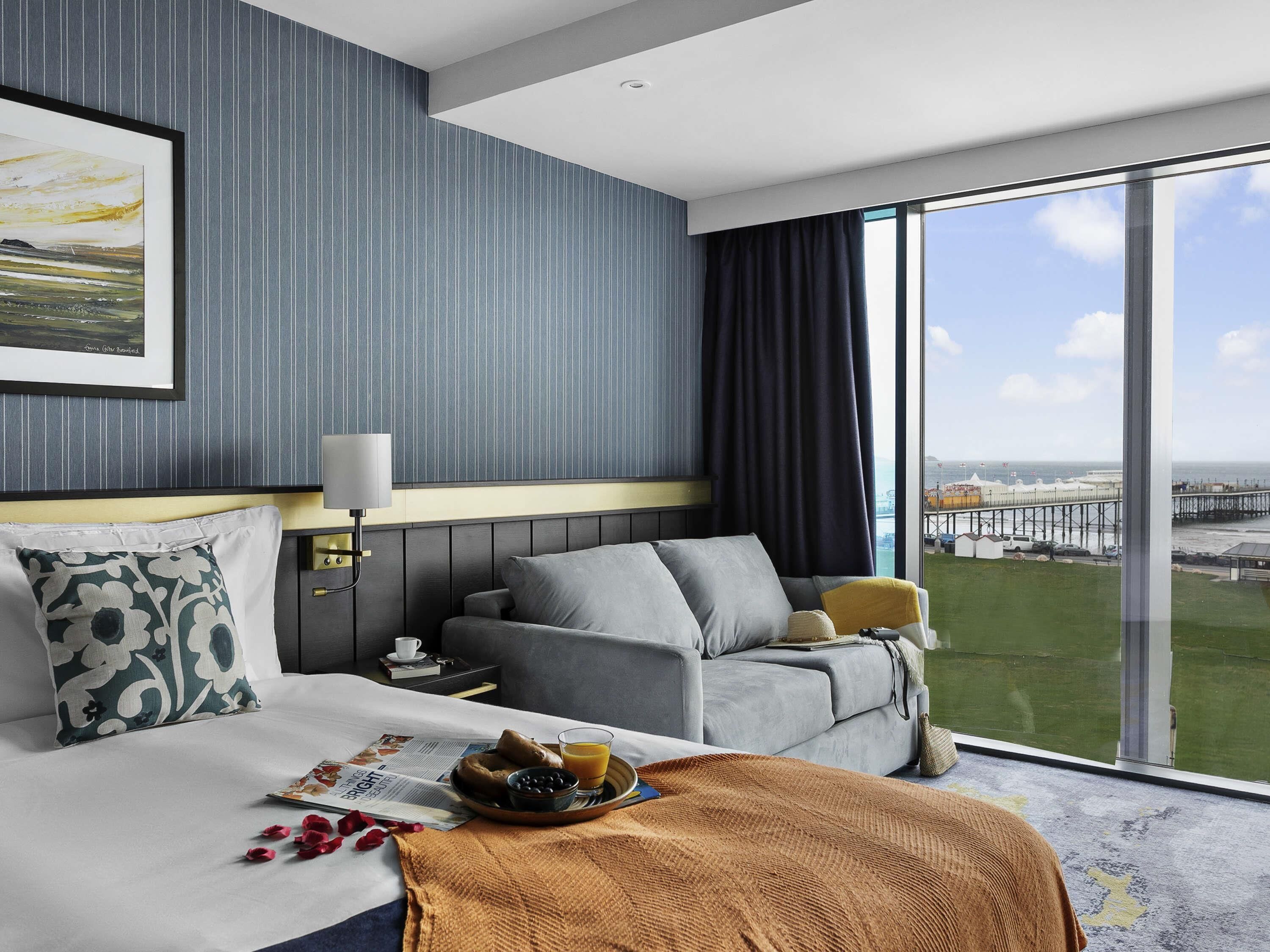 A hotel room with a bed, a sofa, a large window with a view of the sea, and a framed picture on the wall.