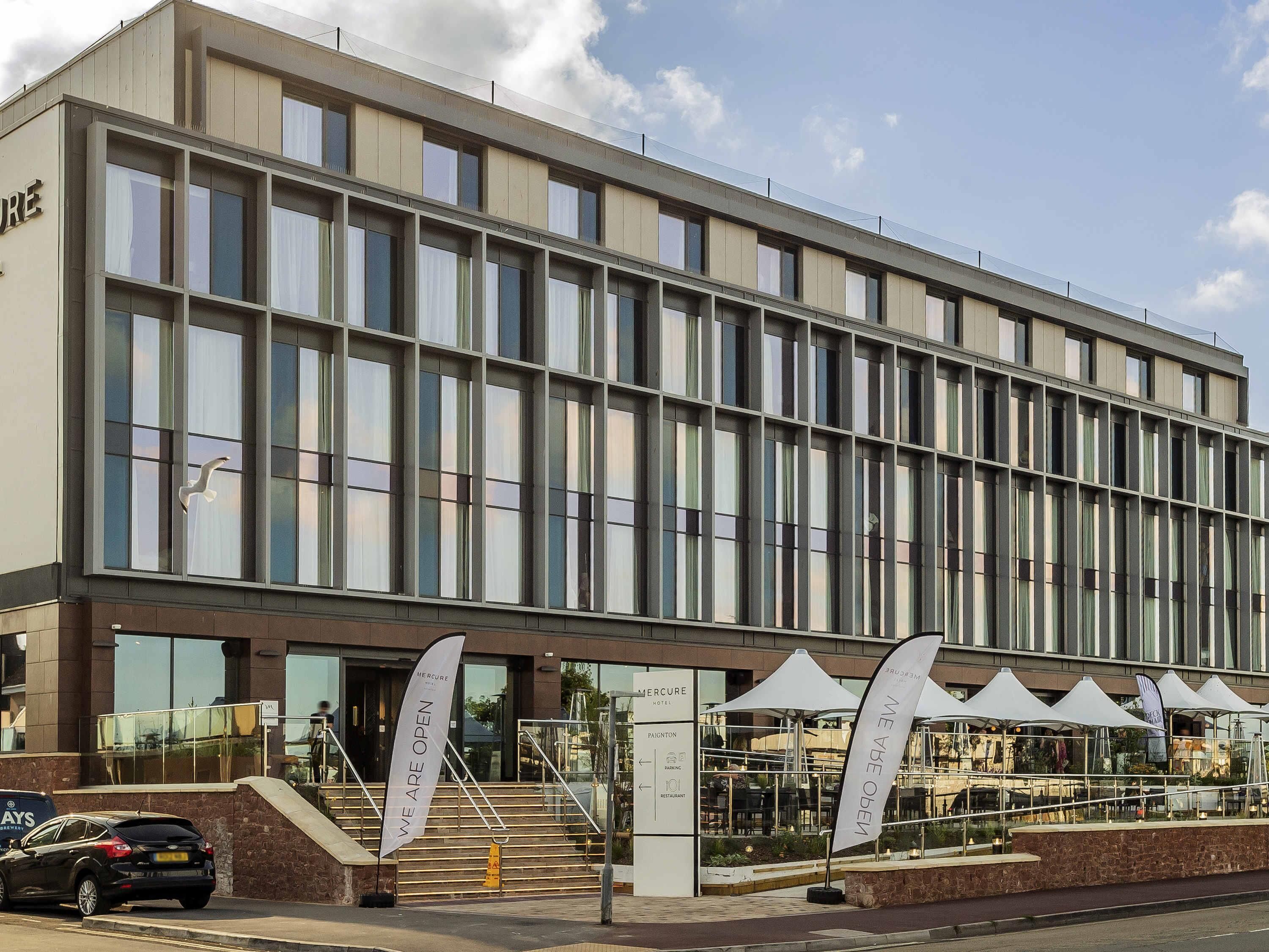 Een modern hotel met een glazen gevel, een duidelijke ingang en een bord waarop staat 'WE ARE OPEN'.