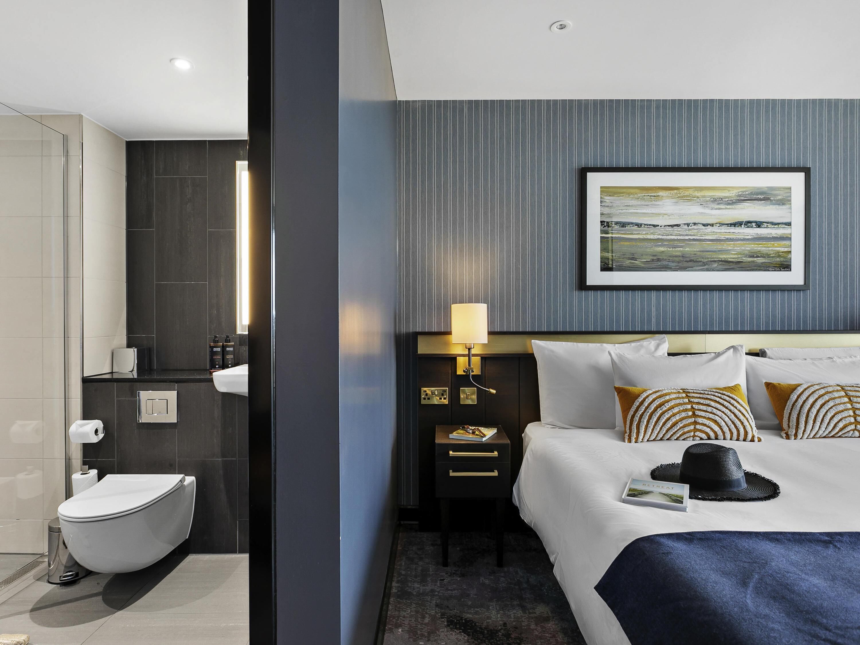 Habitación de hotel con cama, mesita de noche, lámpara, un cuadro en la pared y baño con inodoro y lavabo.