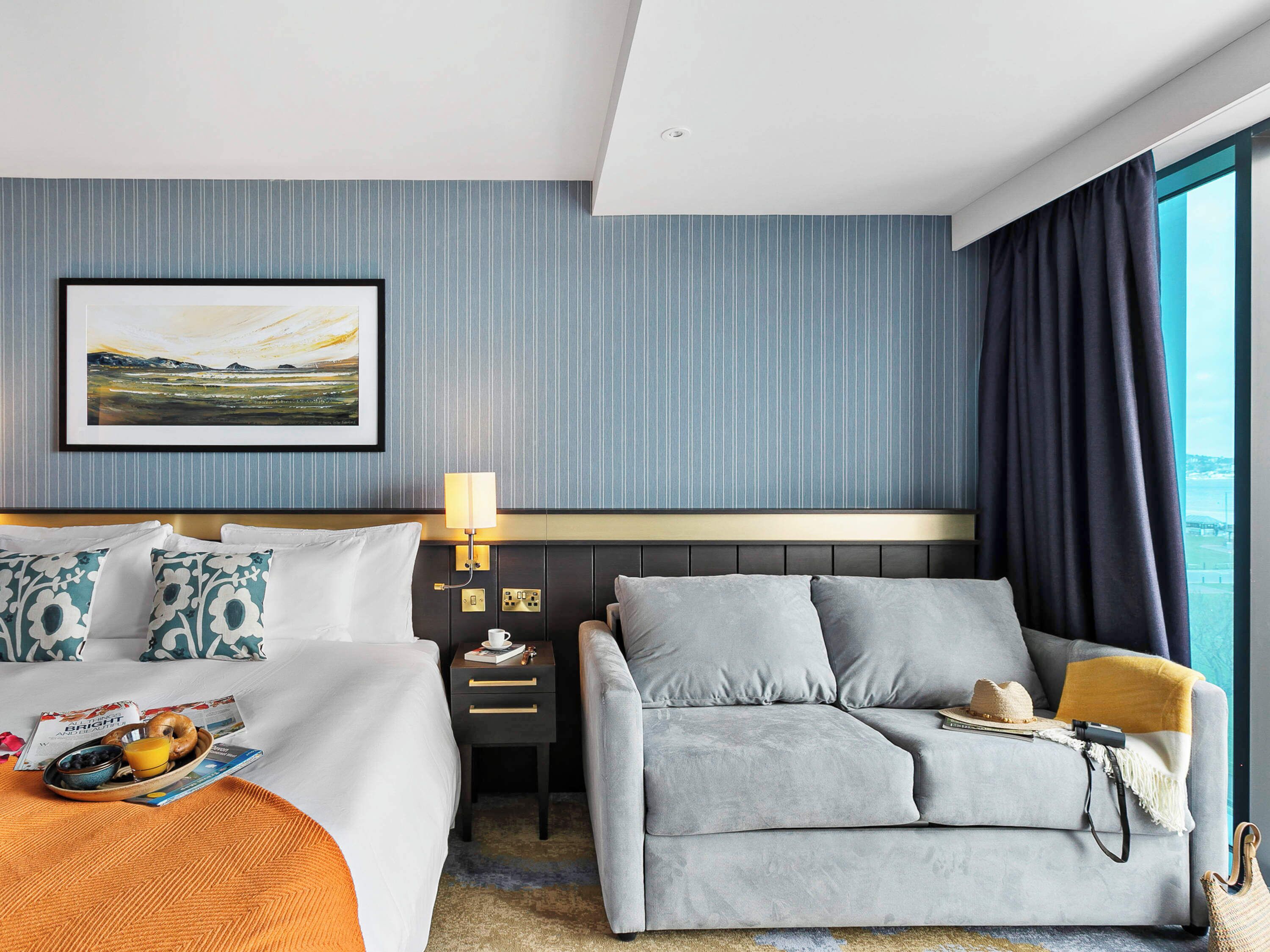 A hotel room with a bed, a sofa, a bedside table with a lamp, a framed picture on the wall, and a window with curtains.
