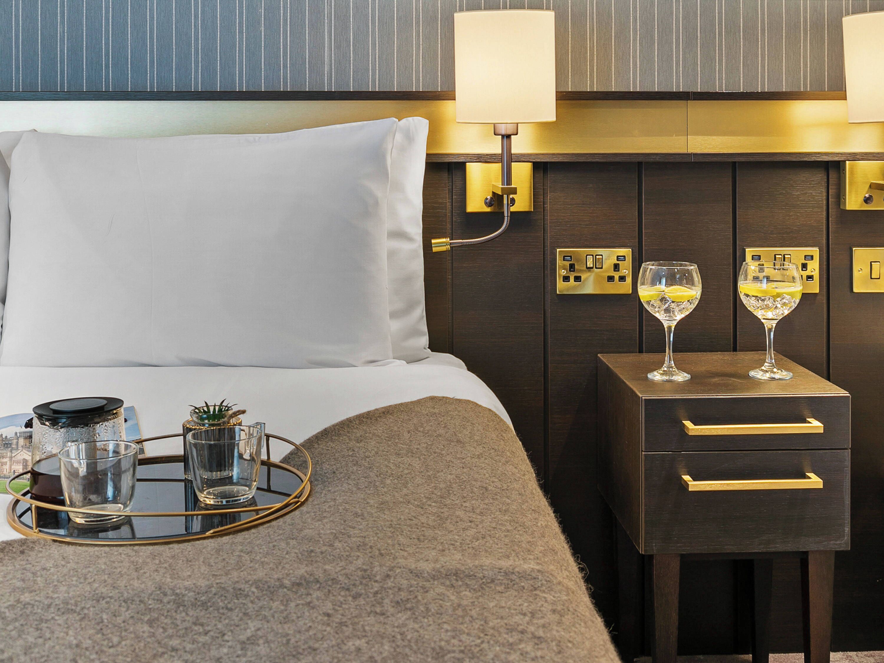 Habitación de hotel con cama, lámparas de noche, una mesita de noche con dos copas de vino y una bandeja con copas y una botella.