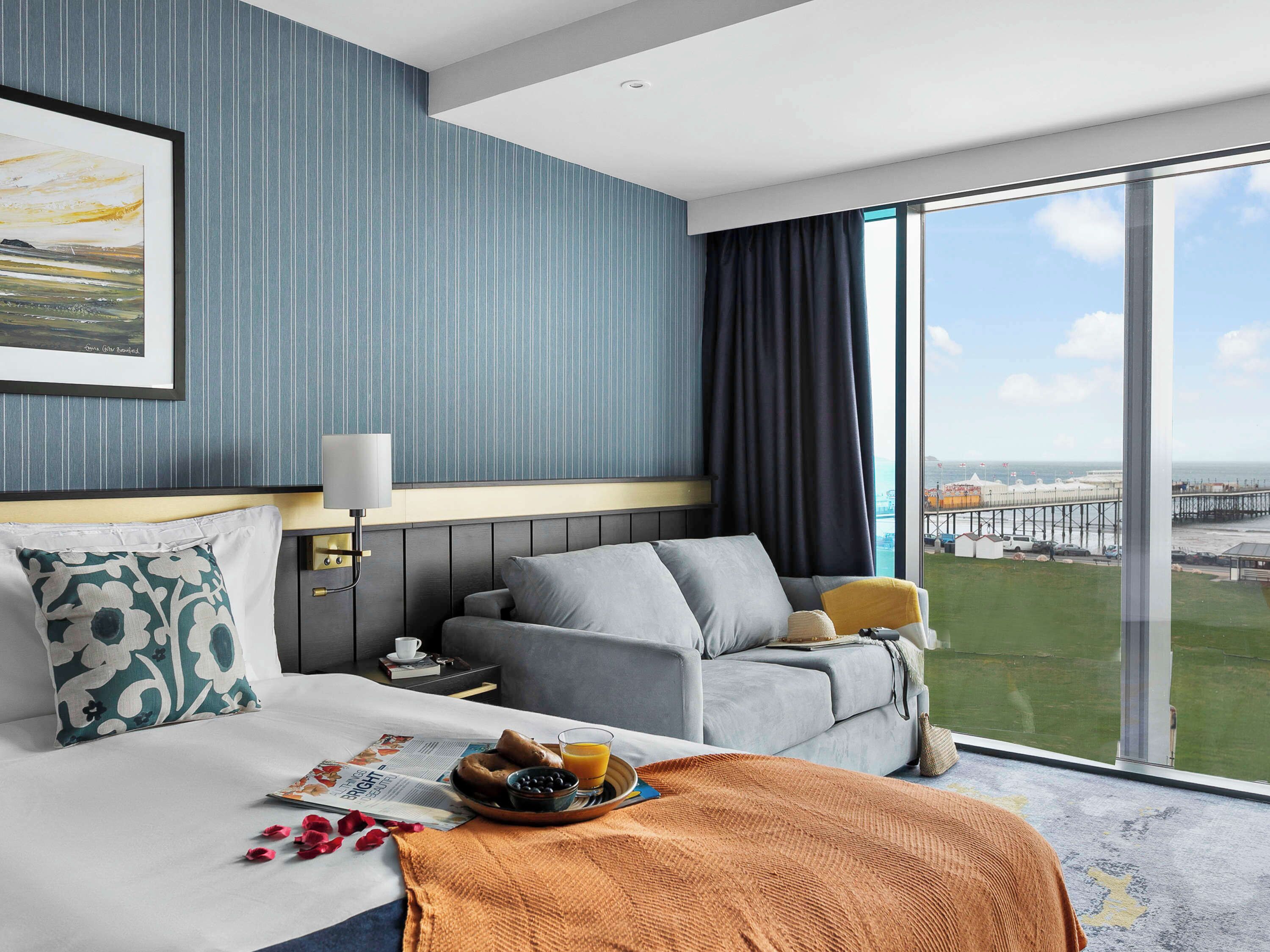 A hotel room with a bed, a sofa, a dining table with breakfast, a large window with a view of the sea, and a framed picture on the wall.