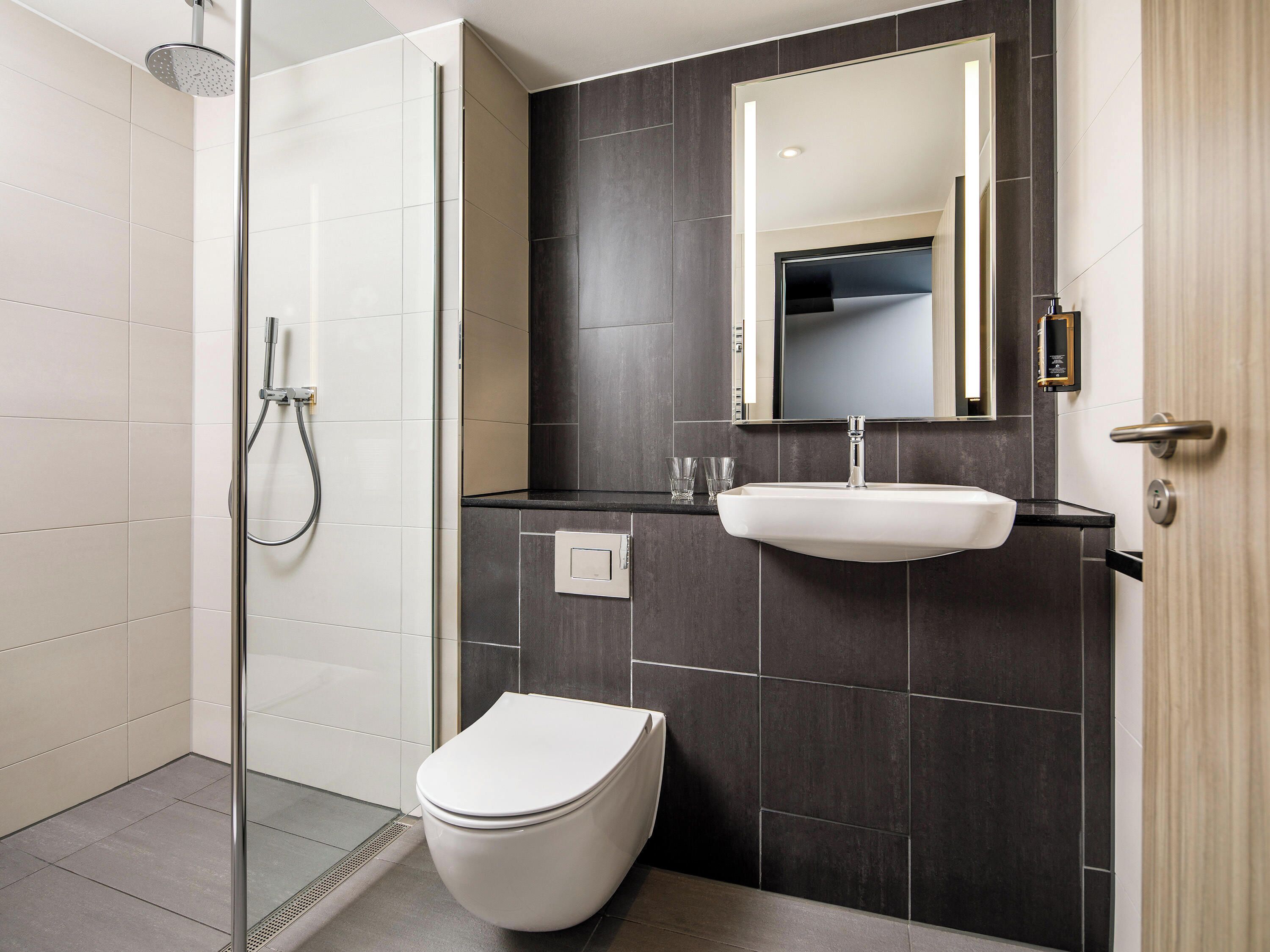 A modern bathroom with a glass-enclosed shower, a white toilet, a sink with a mirror above, and dark tiled walls.