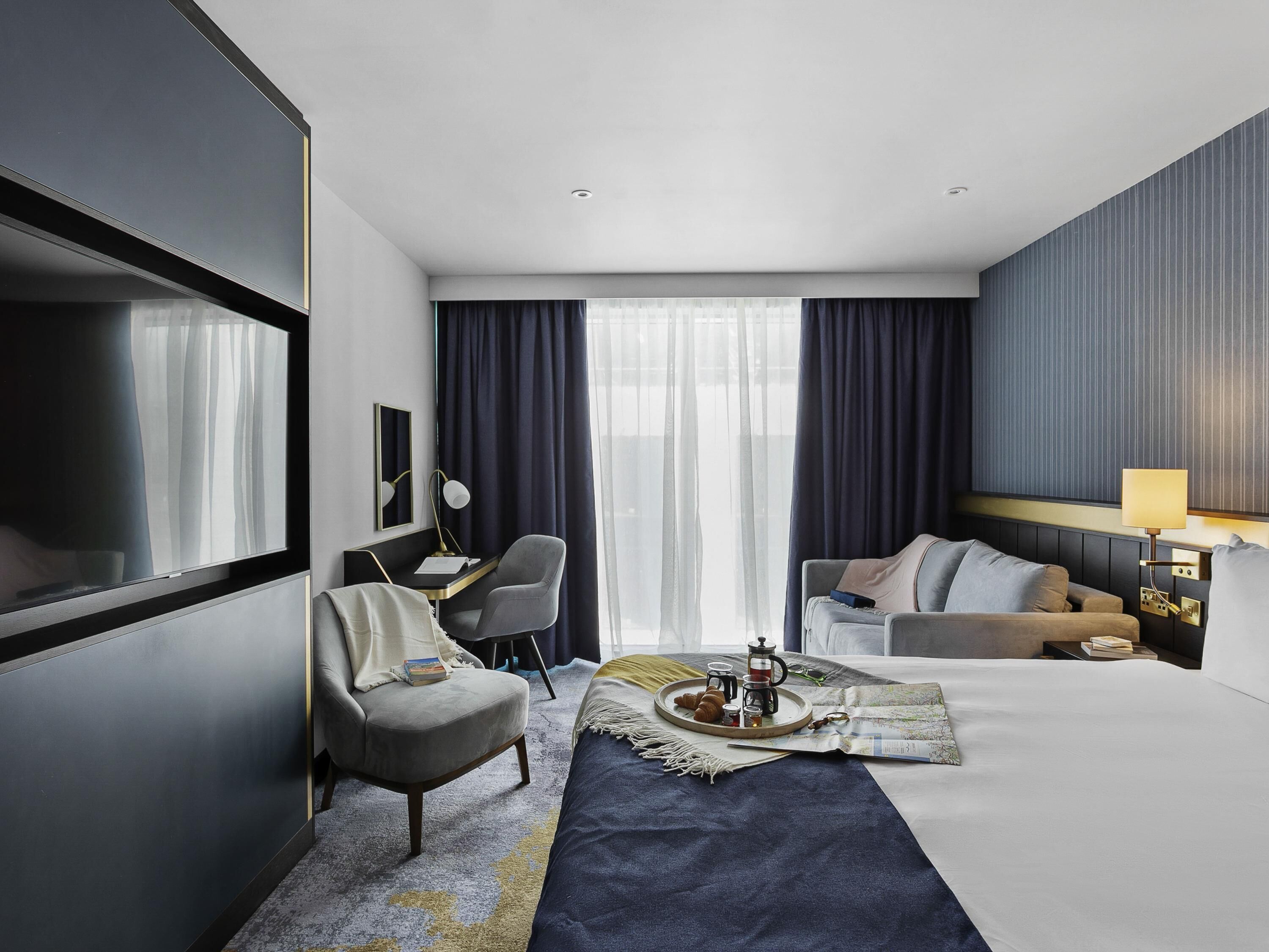 Habitación de hotel con cama, escritorio, silla, sofá, televisor y ventana con cortinas.