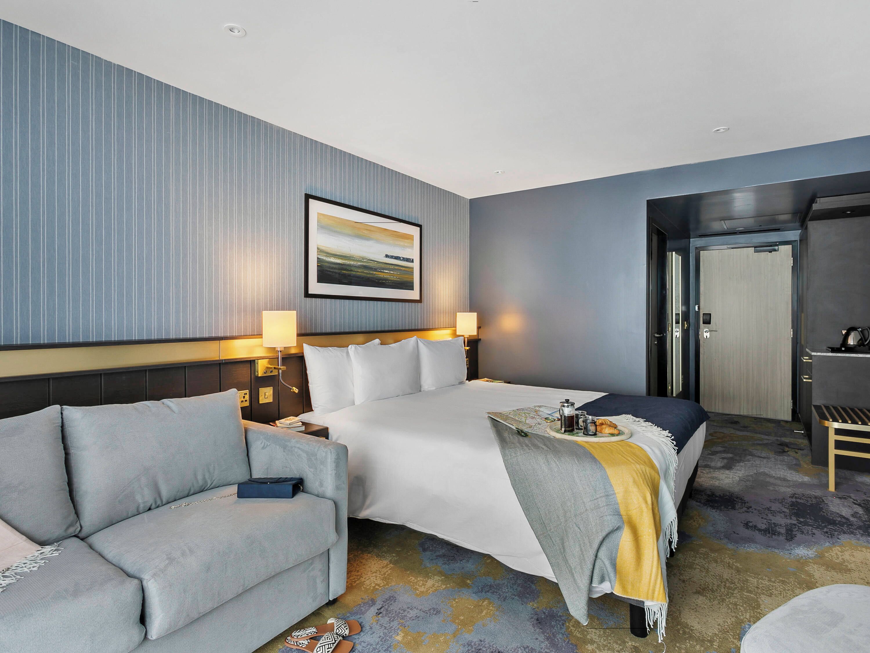 A hotel room with a large bed, a sofa, a desk, and a door.