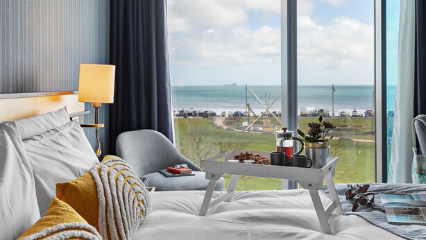 Quarto de hotel com uma grande janela que oferece vista para o mar e uma ilha distante. Há uma cama com roupa de cama branca, uma poltrona cinza e uma pequena mesa com uma bandeja de itens de café da manhã.