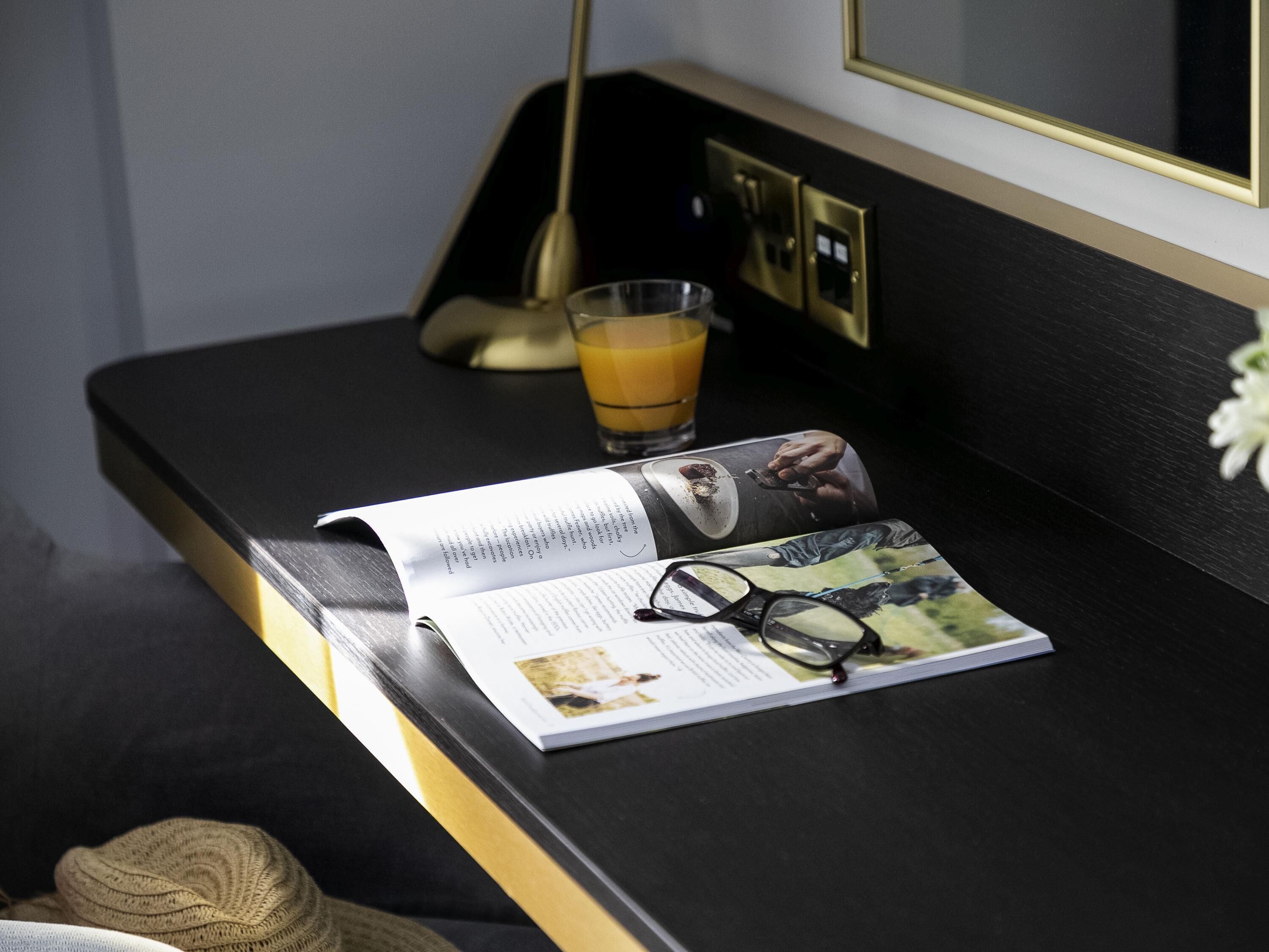 A black table with a magazine, a pair of glasses, and a glass of orange juice.