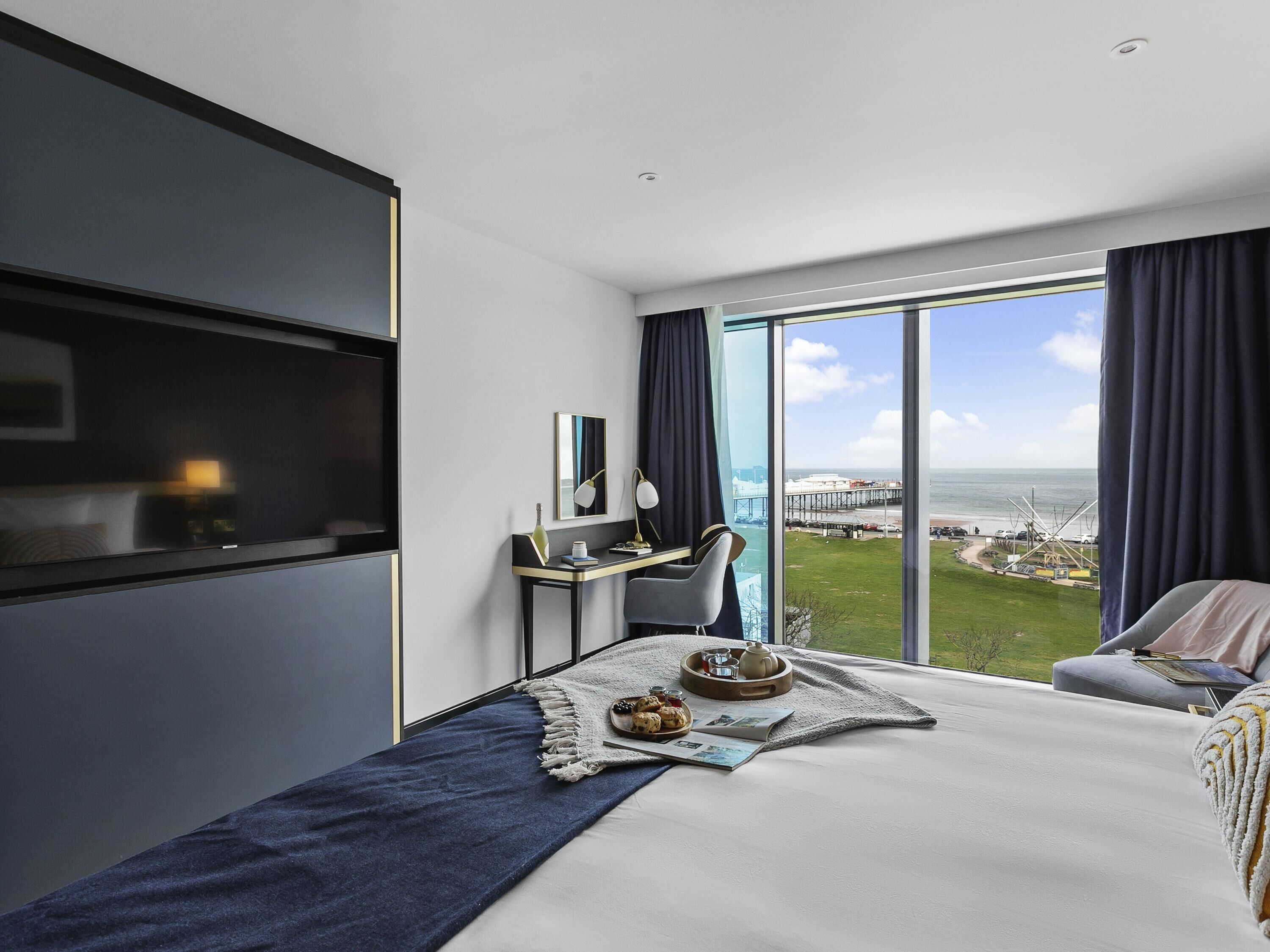 A modern hotel room with a bed, a desk, a chair, and a large window offering a view of the sea and a bridge.