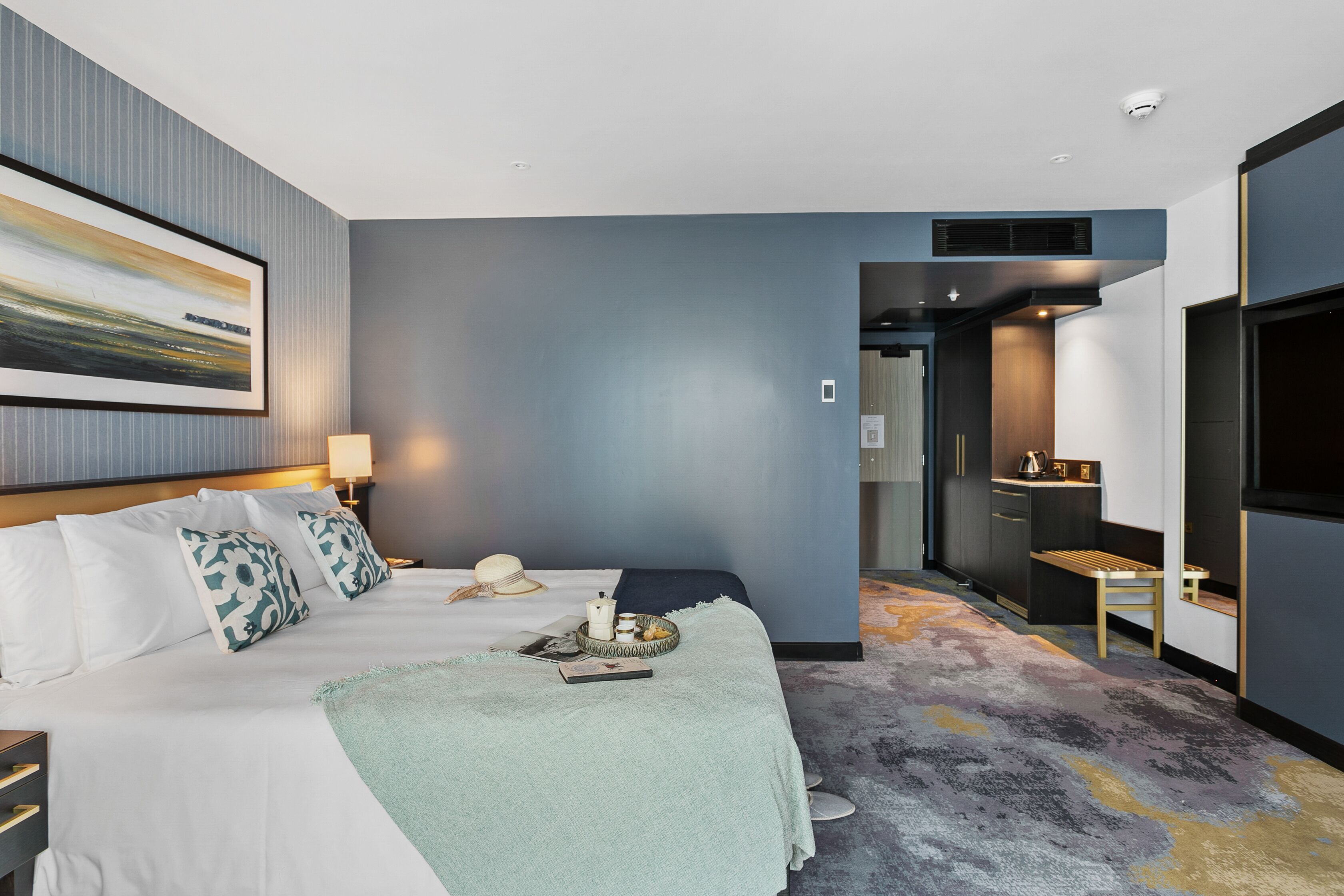 A modern hotel room with a large bed, a desk, and a view of the hallway.