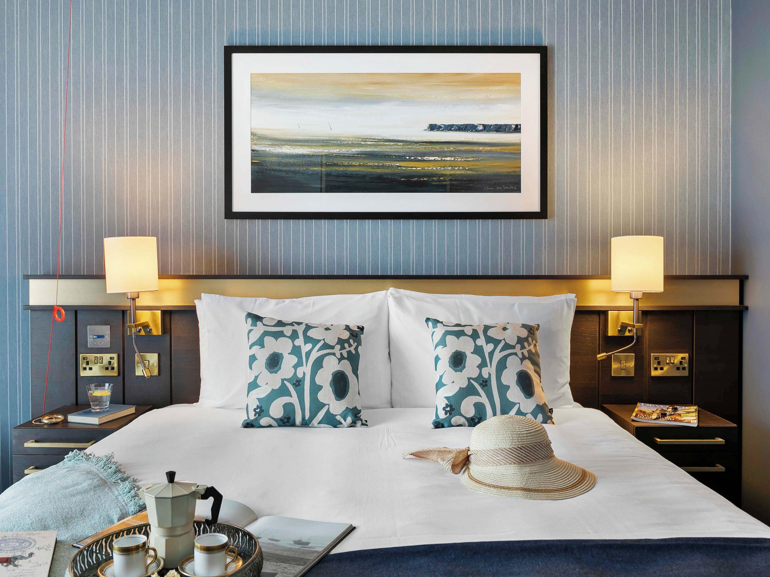 A neatly made bed with white and teal patterned pillows, a straw hat, and a framed landscape painting above it.