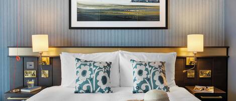 Uma cama bem arrumada com almofadas brancas e com padrão azul-esverdeado, um chapéu de palha e um quadro de paisagem em cima.