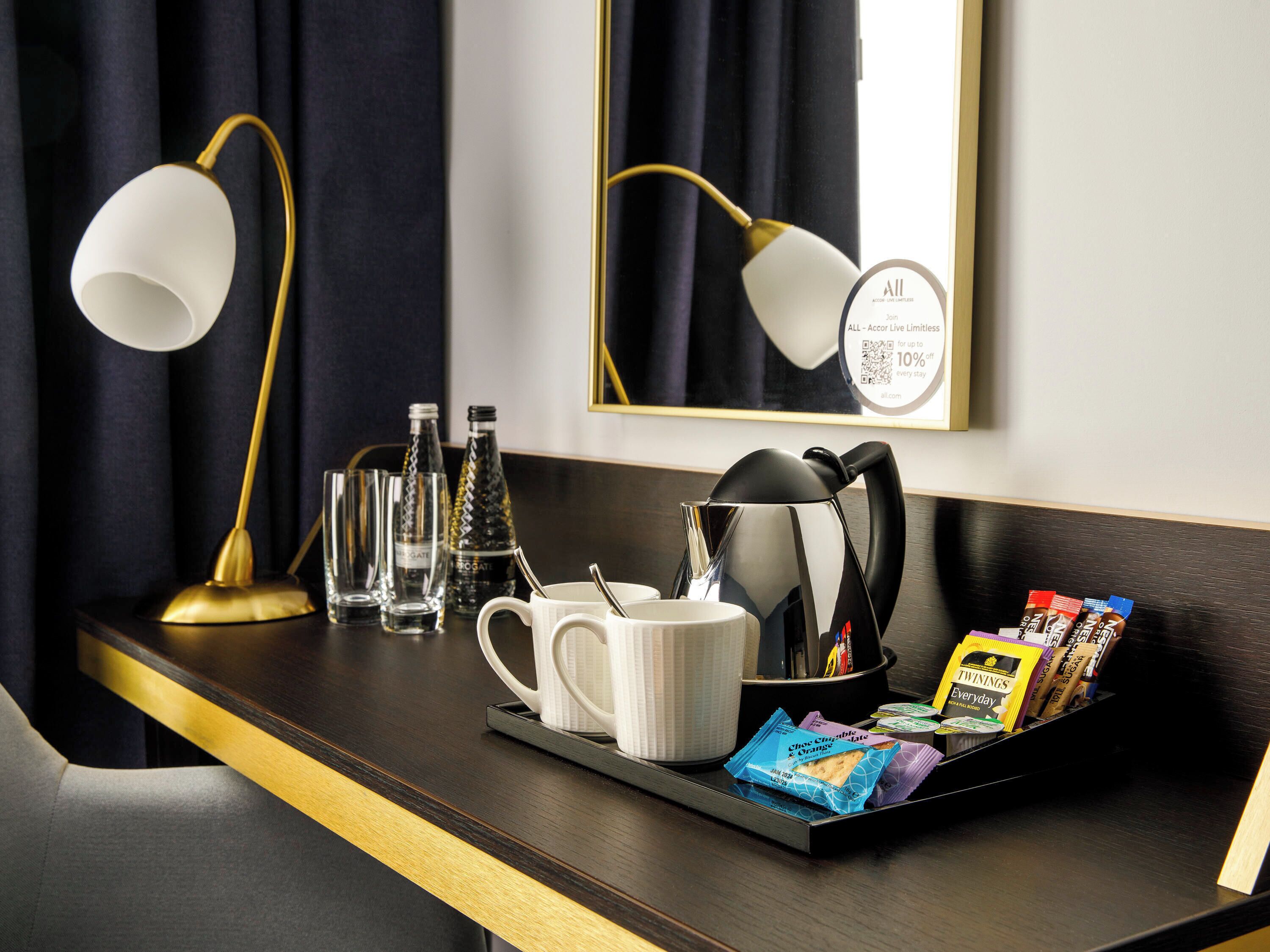 A hotel room with a dark wooden desk, two gold lamps, a white mug, a kettle, and a tray with Twinings tea bags.