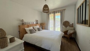 A bedroom with a large bed, wooden headboard, wicker baskets, a wicker chair, and a window with curtains. - Arcachon house 900m from the beach. (Andernos-les-Bains)