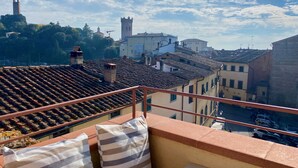 Un balcón con cojines a rayas, techo de baldosas y vista a edificios históricos y una torre al fondo.