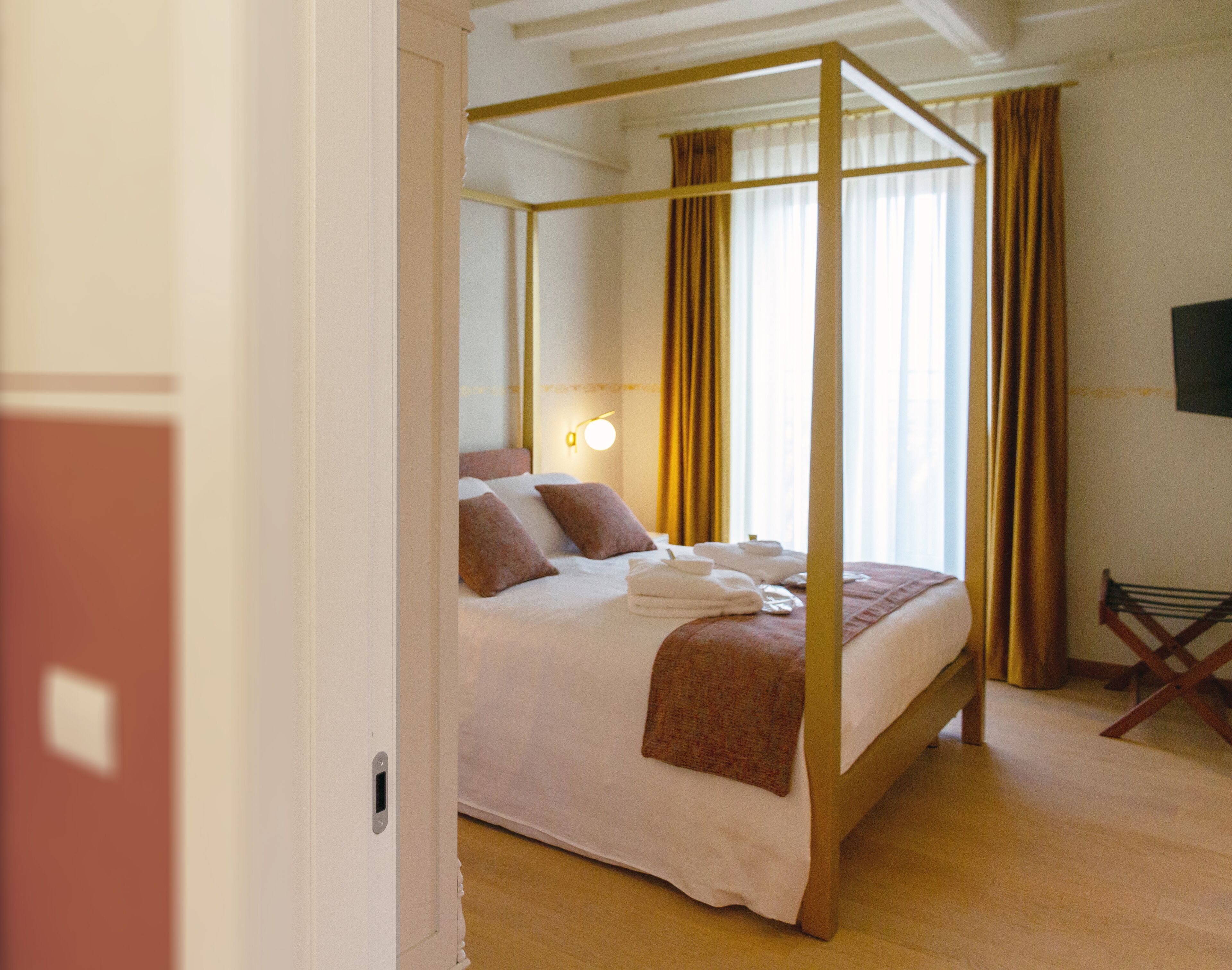 Ein Bett mit Baldachin, ausgestattet mit weißen Leinen und braunen Kissen, ein Nachttisch mit Lampe sowie ein Sessel in einem Hotelzimmer.