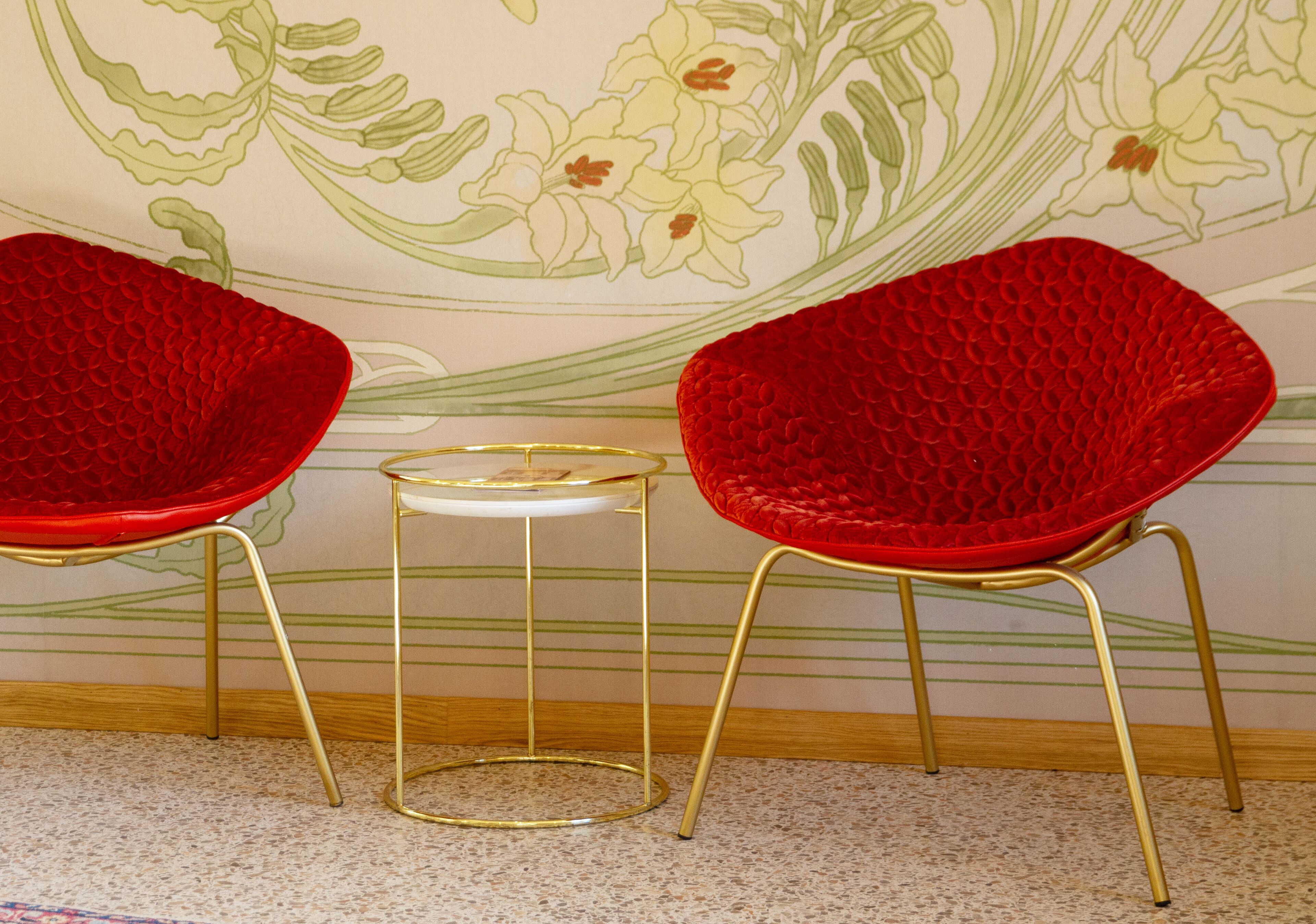 Two red chairs with gold legs and a small white table with a gold base, positioned in front of a floral wallpaper.