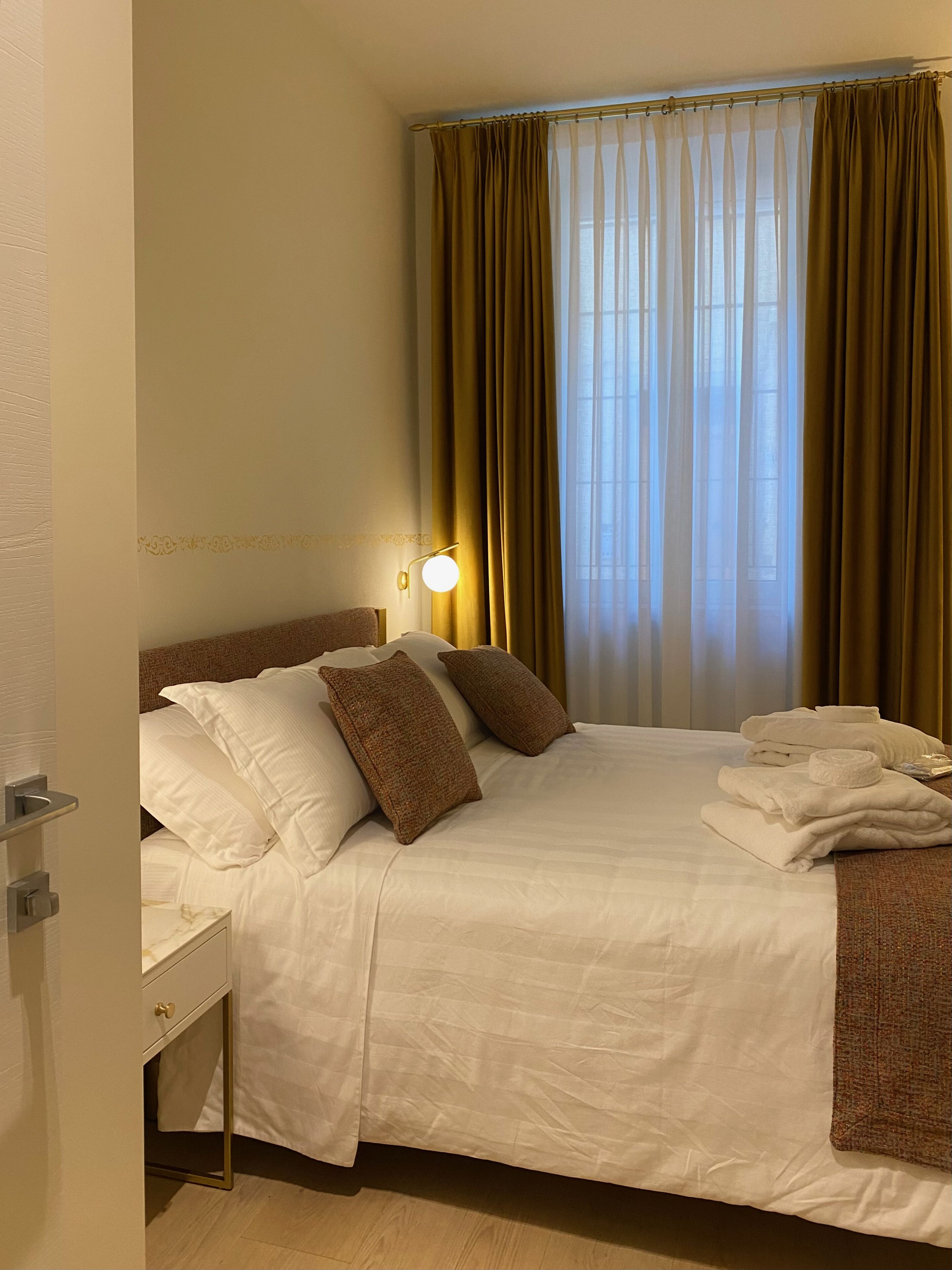 Una cama bien hecha con ropa de cama blanca y marrón, una lámpara de noche y una ventana con cortinas.