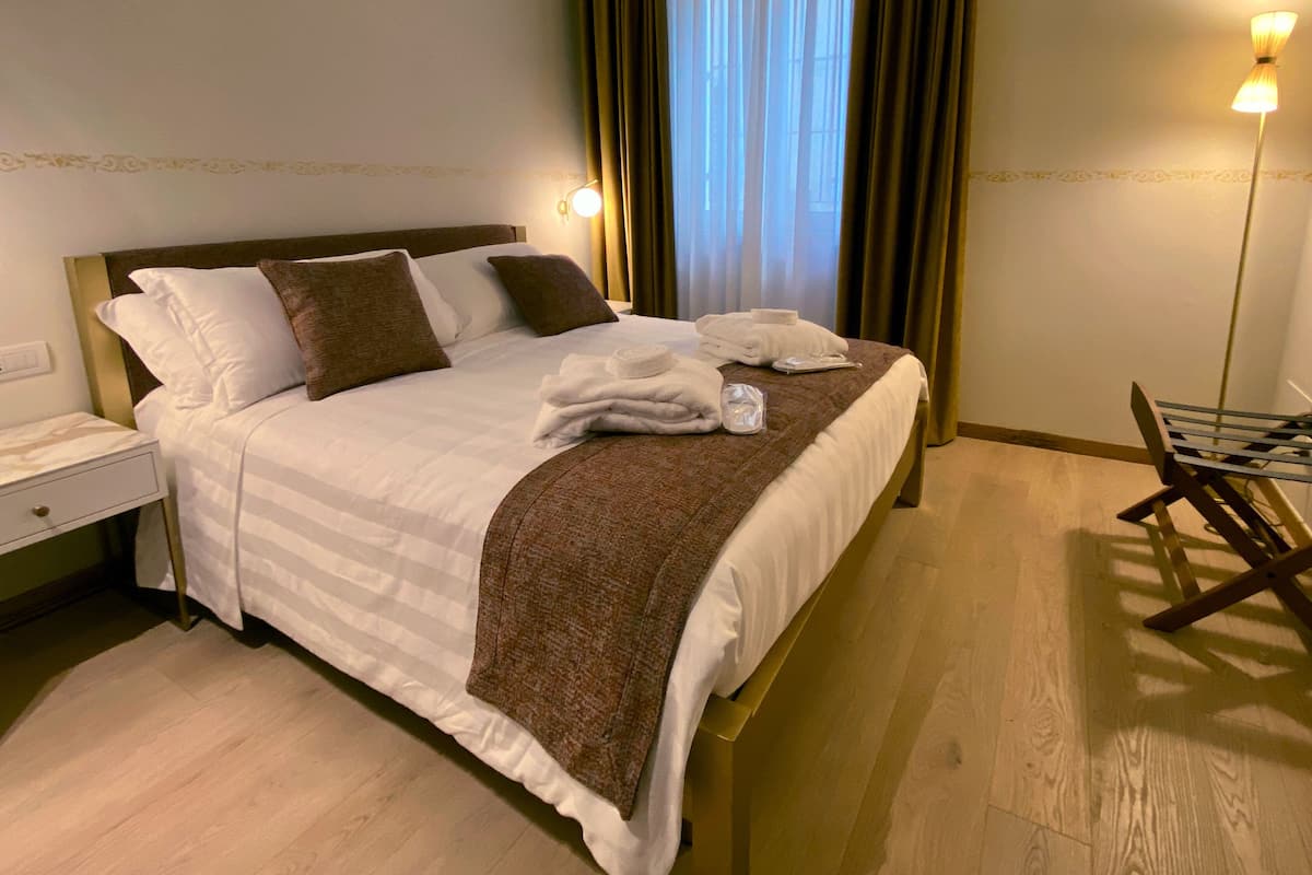 Una cama bien hecha con almohadas, una toalla doblada y una manta. Se ve una mesita de noche con una lámpara y una silla con una bandeja.