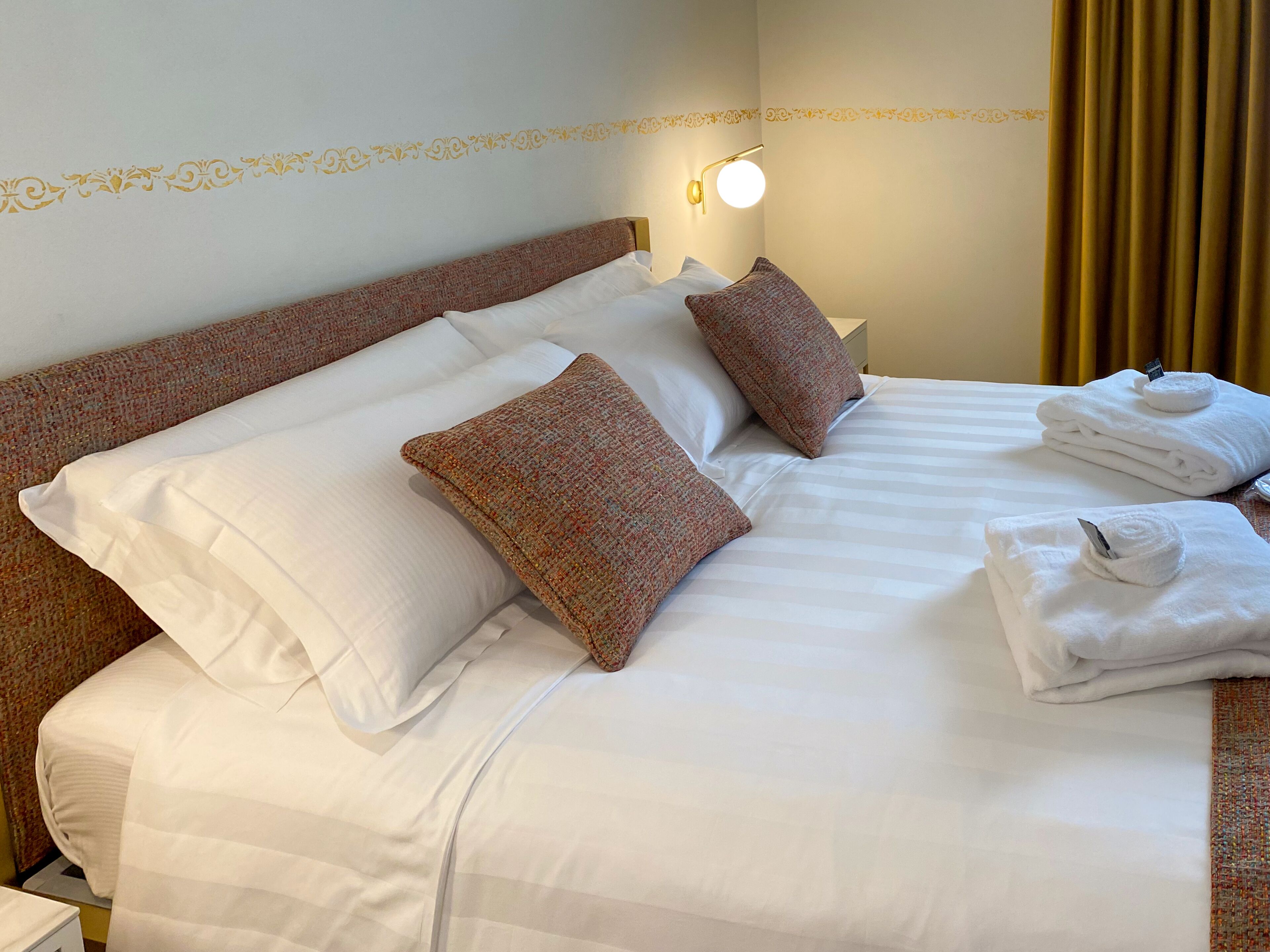 Una cama bien tendida con sábanas blancas, dos almohadas decorativas y un juego de toallas dobladas.