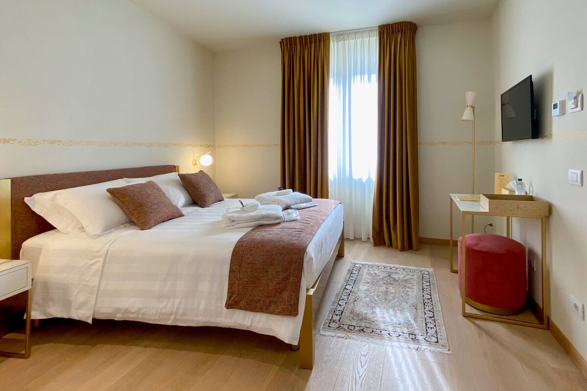 Habitación de hotel con cama, mesita de noche, lámpara, televisor y un otomano rojo.