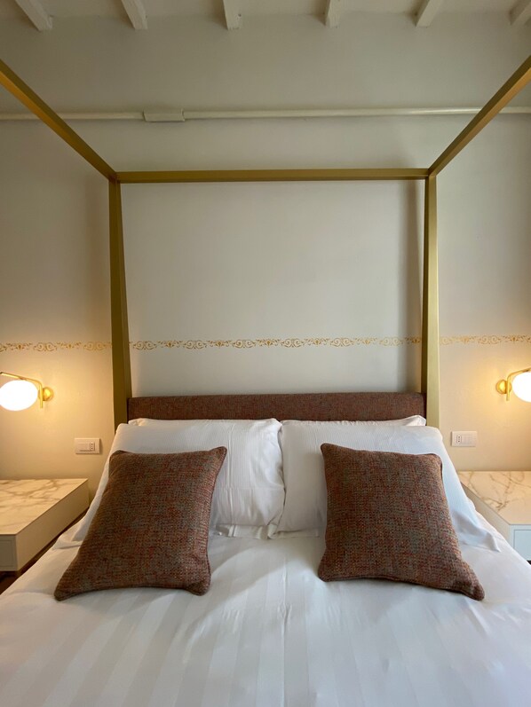 Uma cama bem arrumada com lençóis brancos e duas almofadas decorativas, cabeceira com detalhes ornamentais e luminárias fixadas na parede em ambos os lados.