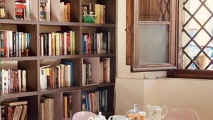 Una habitación con una estantería llena de libros, una mesa dispuesta para el té con sillas rosadas y un ventanal con marco de madera.