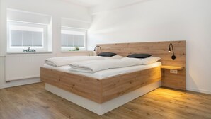 Un dormitorio moderno con cama de madera, ropa de cama blanca, una mesita de noche con lámpara y una ventana con persianas.