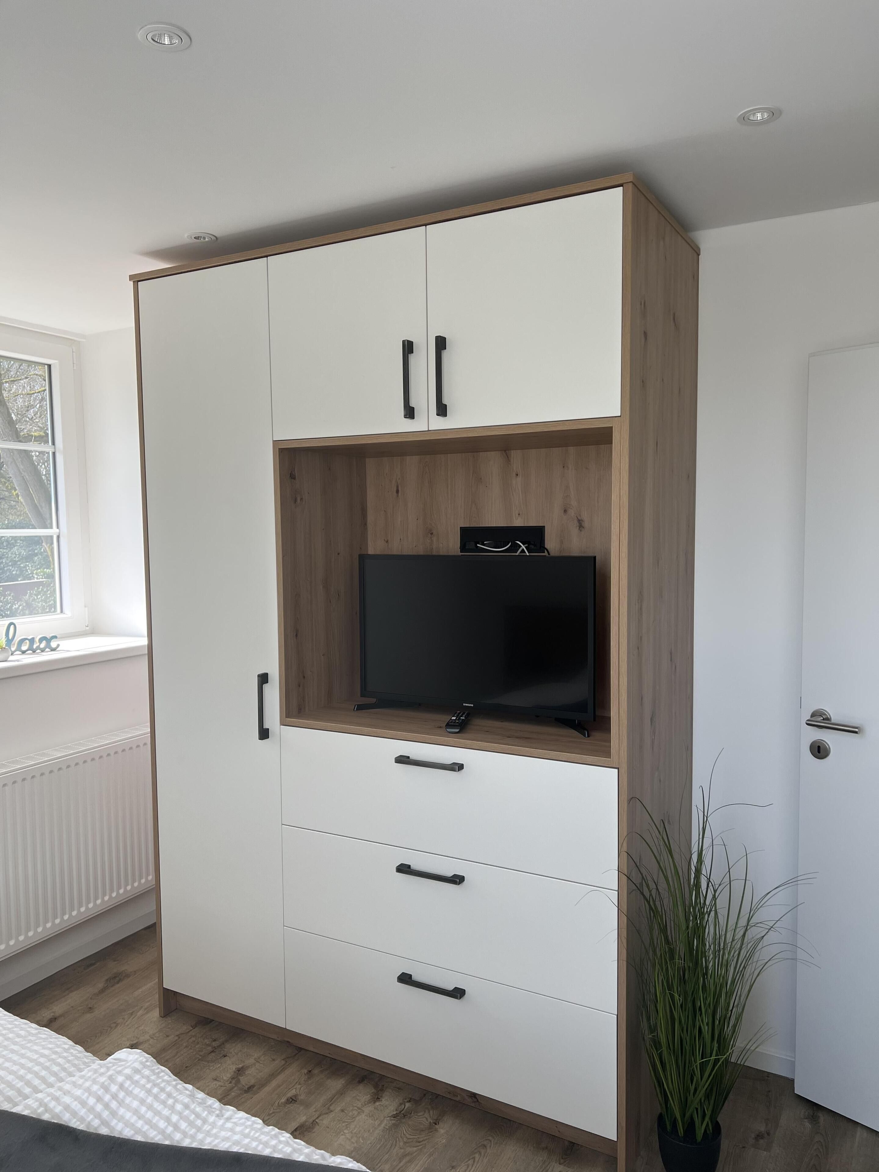 Una habitació amb un armari blanc amb detalls de fusta, un televisor de pantalla plana sobre una prestatgeria de fusta i una planta en test.