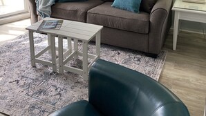 Un salon avec un canapé marron, deux coussins turquoise, une table basse blanche, un fauteuil en cuir noir, un tapis à motifs, un tableau représentant des voiliers et une lampe sur une table d’appoint.