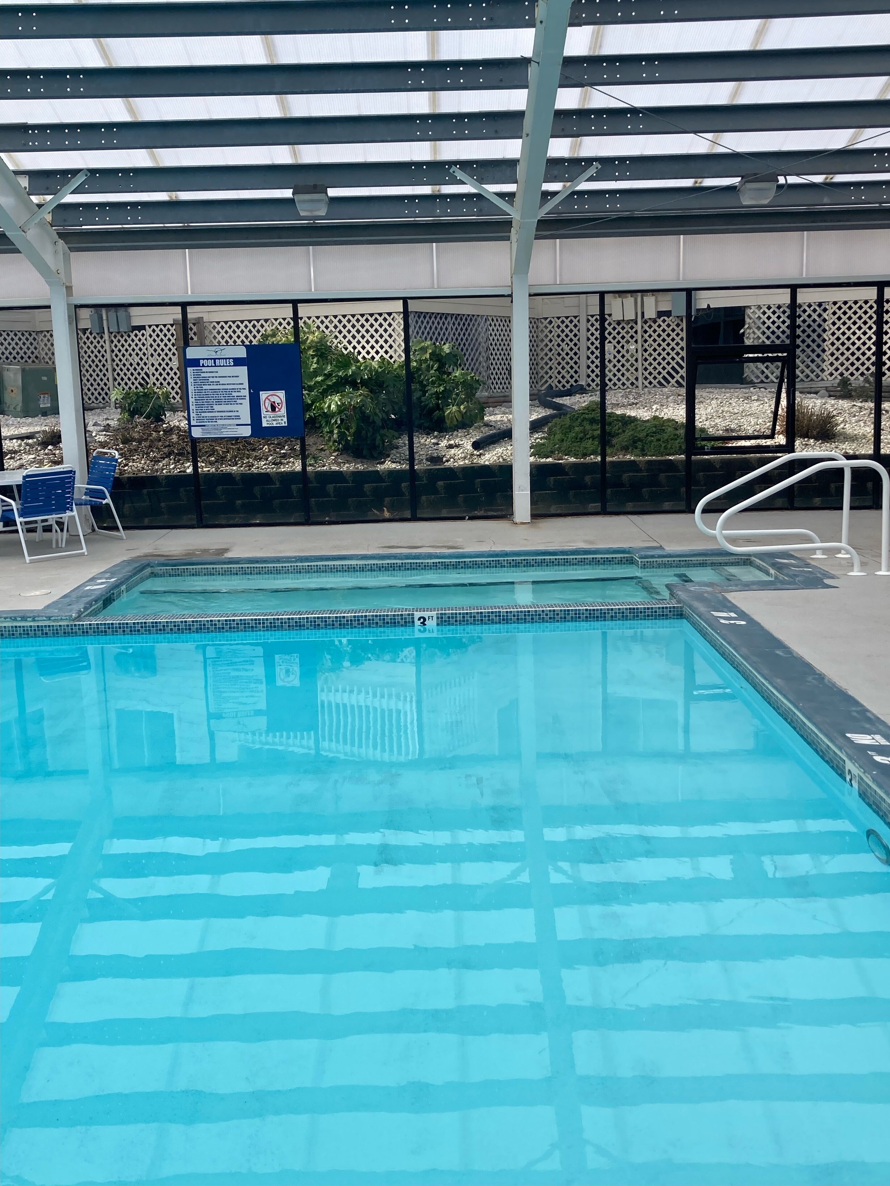 A swimming pool with a covered area, pool rules sign, and blue chairs.