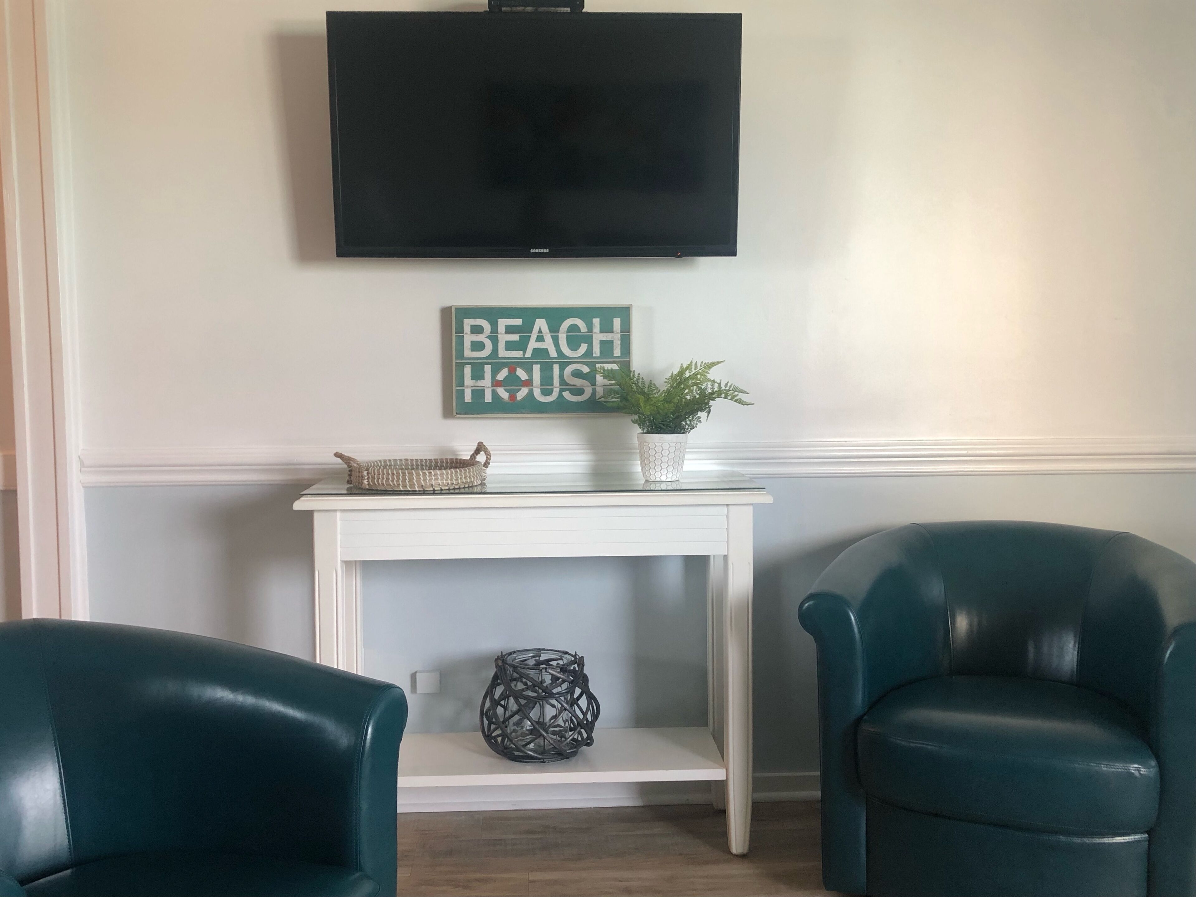 A living room with teal chairs, a white console table, a flat-screen TV, and a 'BEACH HOUSE' sign.