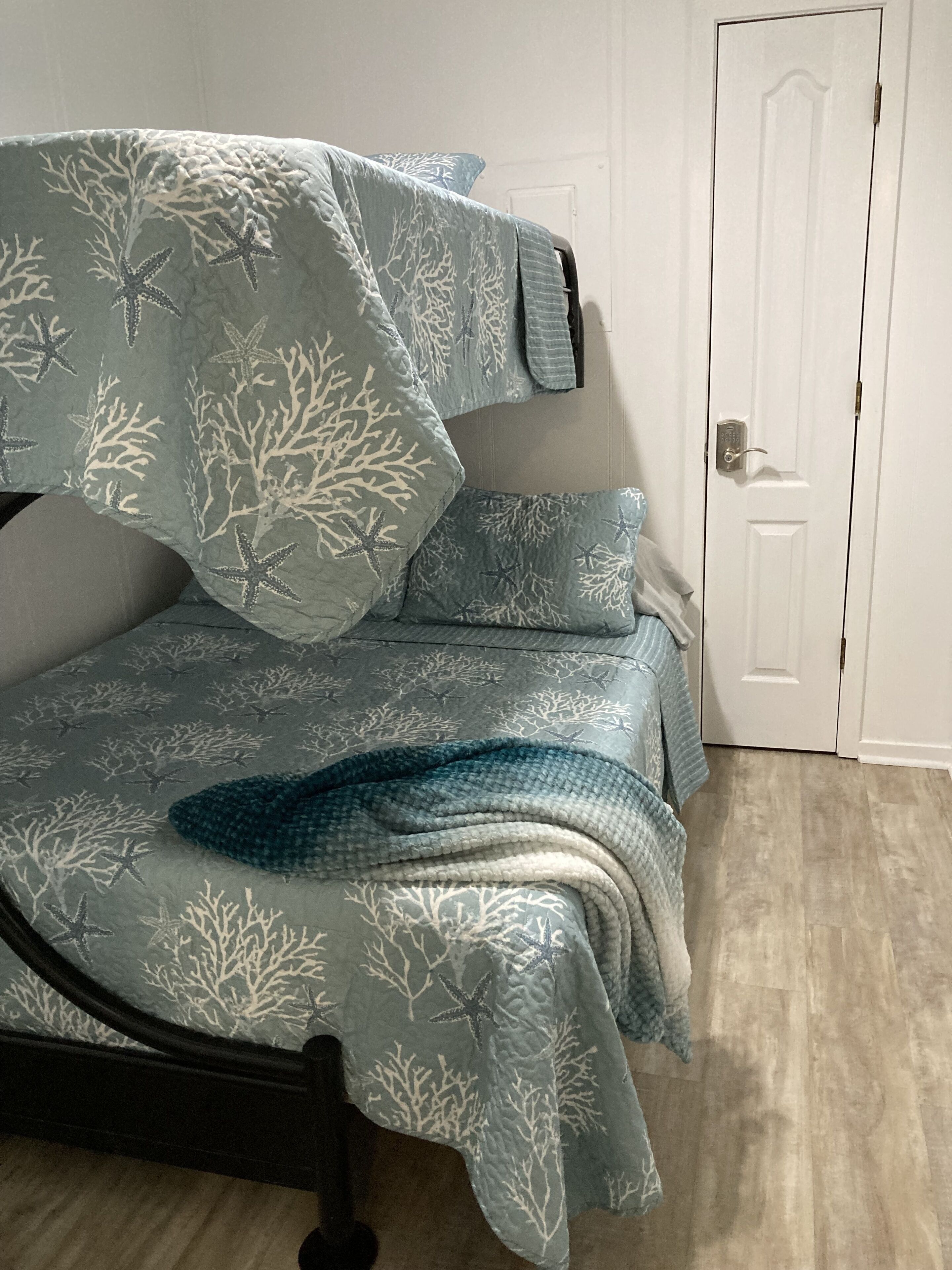 Una cama con colcha y almohadas de estampado coral azul y blanco, un cabecero de madera oscura y una puerta blanca al fondo.