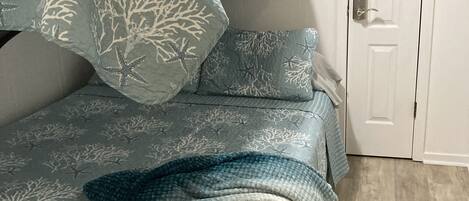 Una cama con colcha y almohadas de estampado coral azul y blanco, un cabecero de madera oscura y una puerta blanca al fondo.