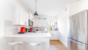 A modern kitchen with white cabinetry, a stainless steel refrigerator, a central island with bar stools, and a pendant light.