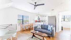 A modern living room with a grey sofa, a wooden coffee table, and a dining area with a table and chairs. There is a kitchen in the background with white cabinets and a refrigerator.