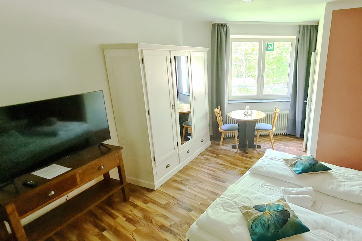 A hotel room with a TV, a wardrobe, a bed with pillows, a small table, and a chair.
