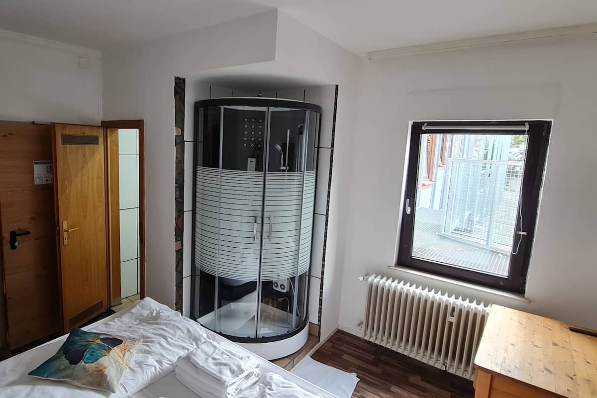 A room with a bed, a wooden wardrobe, a bathroom with a glass-enclosed shower, a window with a view, and a radiator.